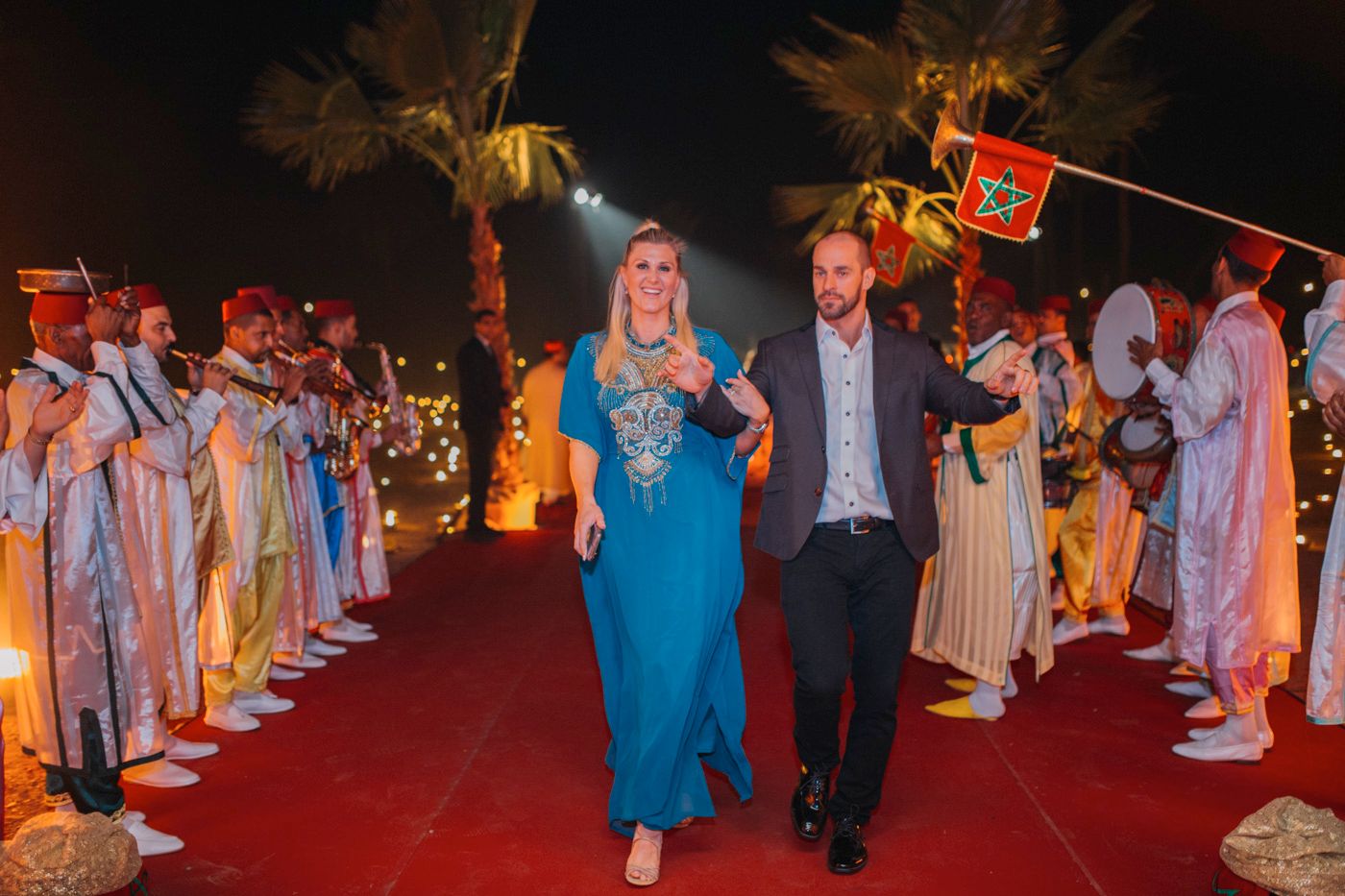 Guests in traditional dress proceed down a red carpet lined with performers in matching attire.