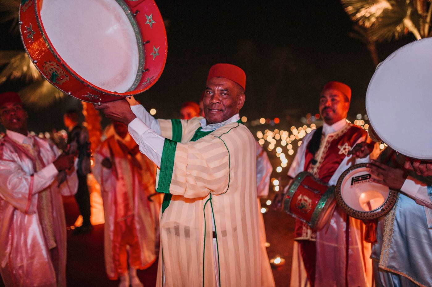 Traditional Moroccan musicians in red fez hats perform with large drums during a nighttime celebration.