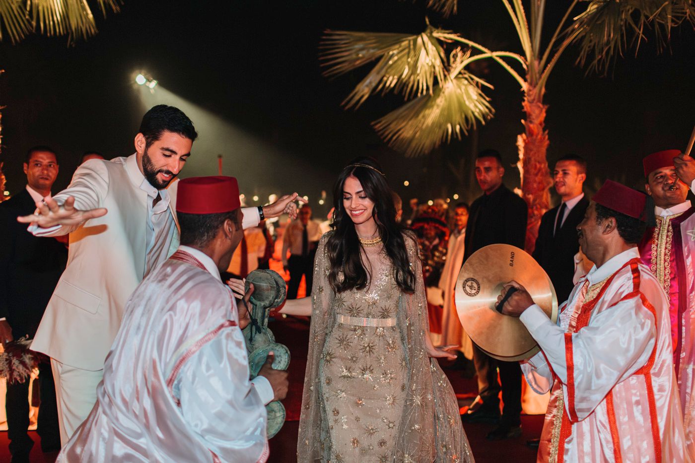 Wedding celebration with traditional Moroccan musicians and dancers under palm trees at night.