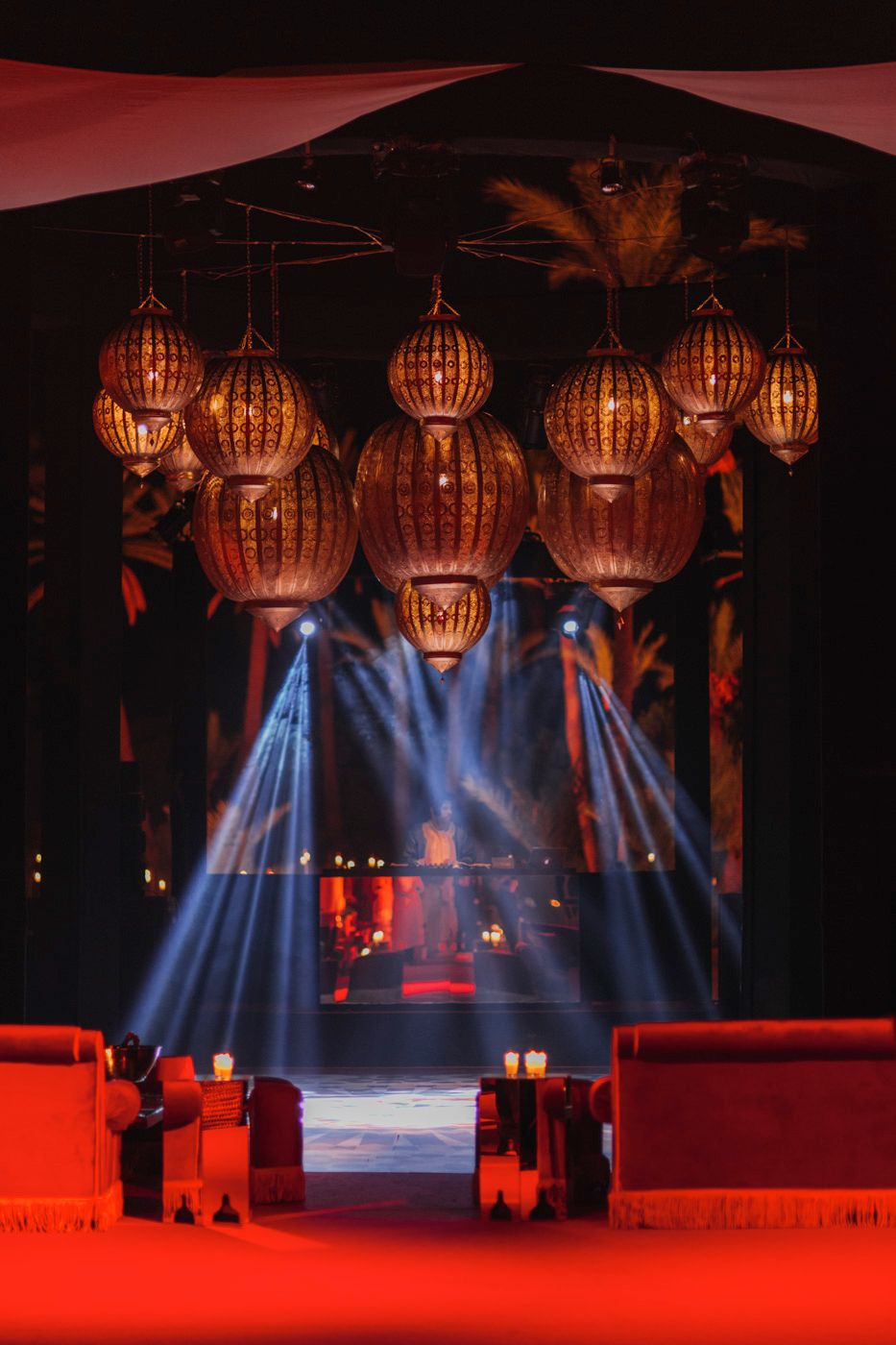 Ornate Moroccan lanterns hanging above red-lit walkway with dramatic blue spotlights.