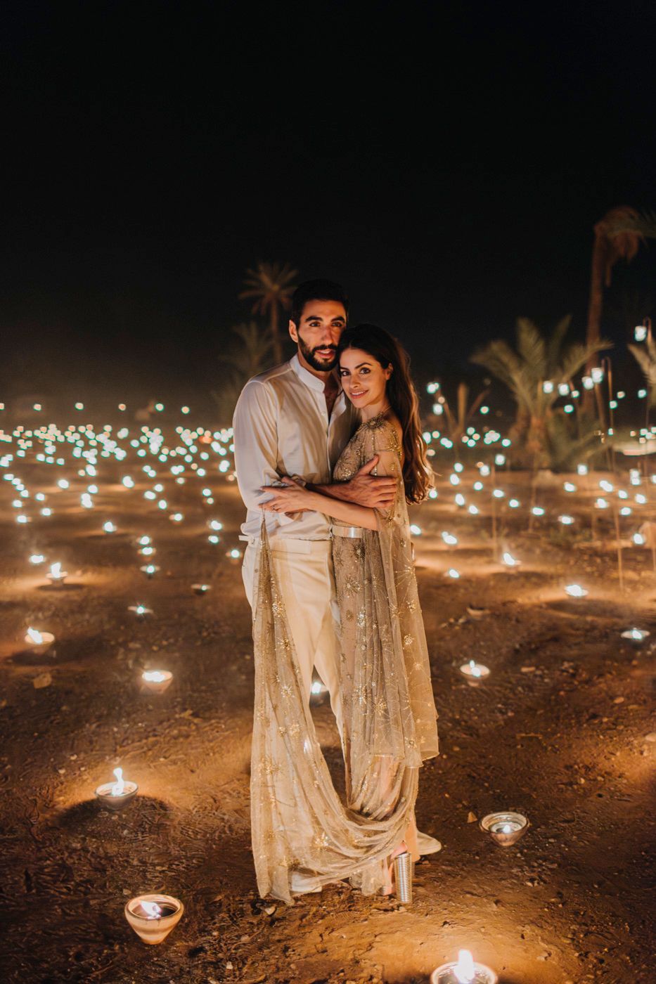 Romantic couple embracing under twinkling lights and palm trees in an evening desert setting.