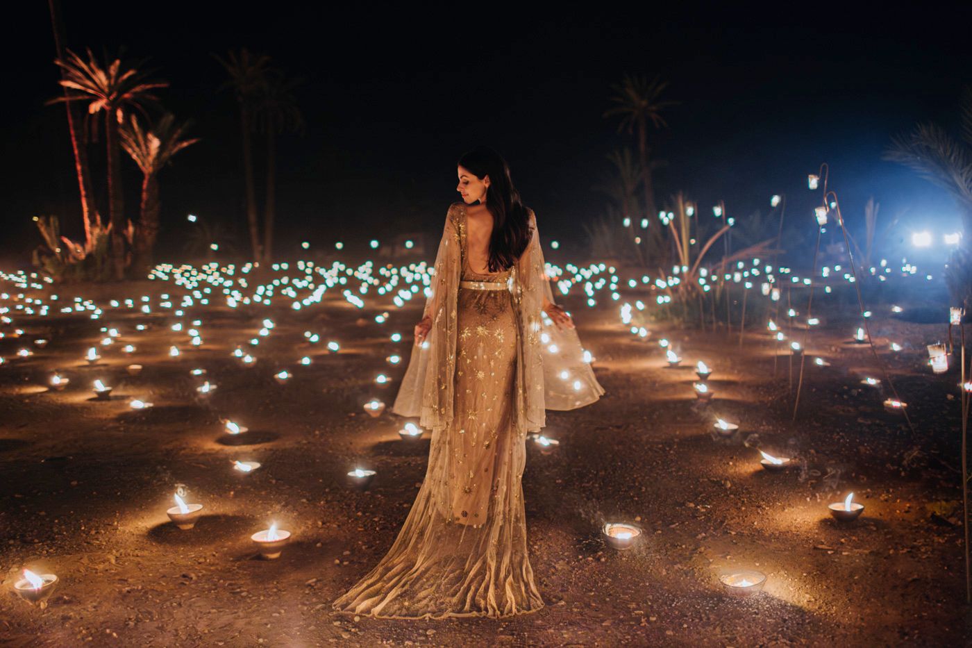 Person in a shimmering gown stands among scattered ground lights and palm trees.
