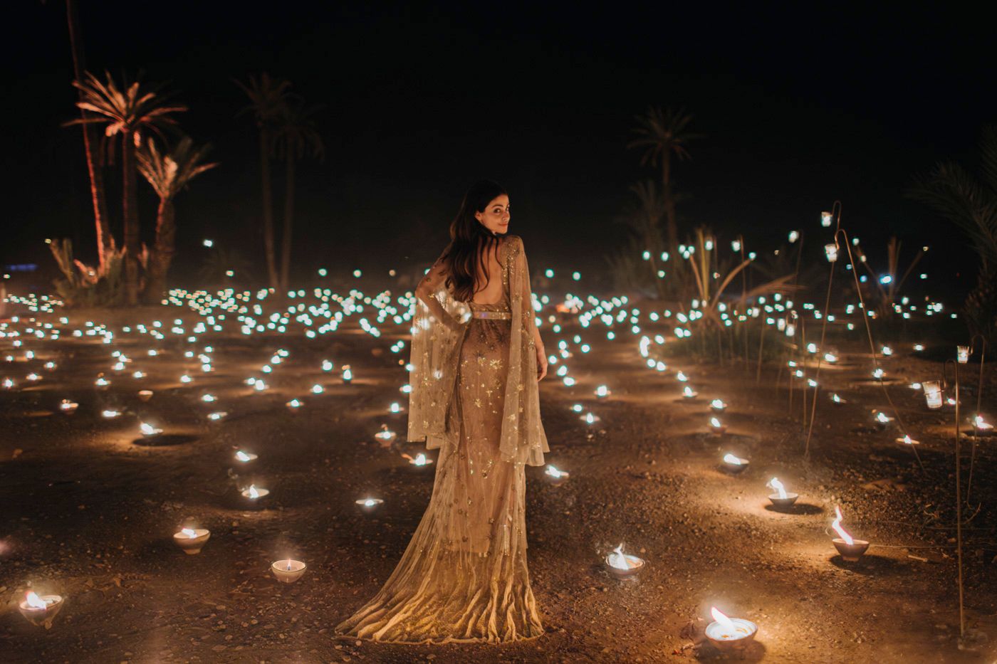 Figure in a golden dress photographed among scattered lights in a palm grove.