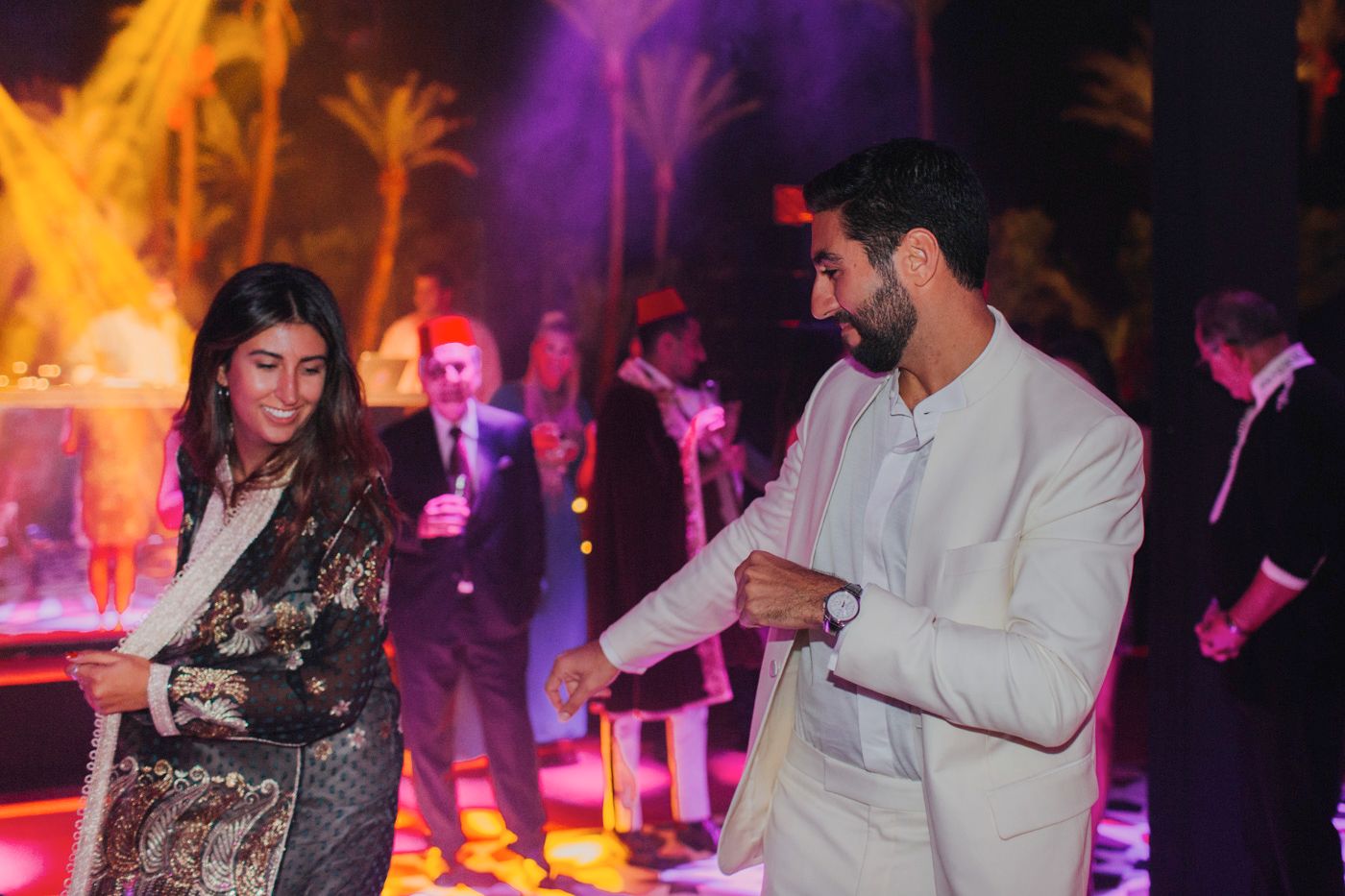 Couple dancing at an evening outdoor party with colorful lighting and palm trees.