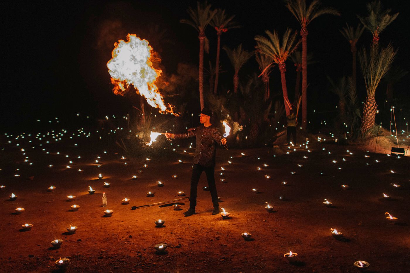 Fire performer creating dramatic flame effects against a palm tree backdrop with ground lighting.
