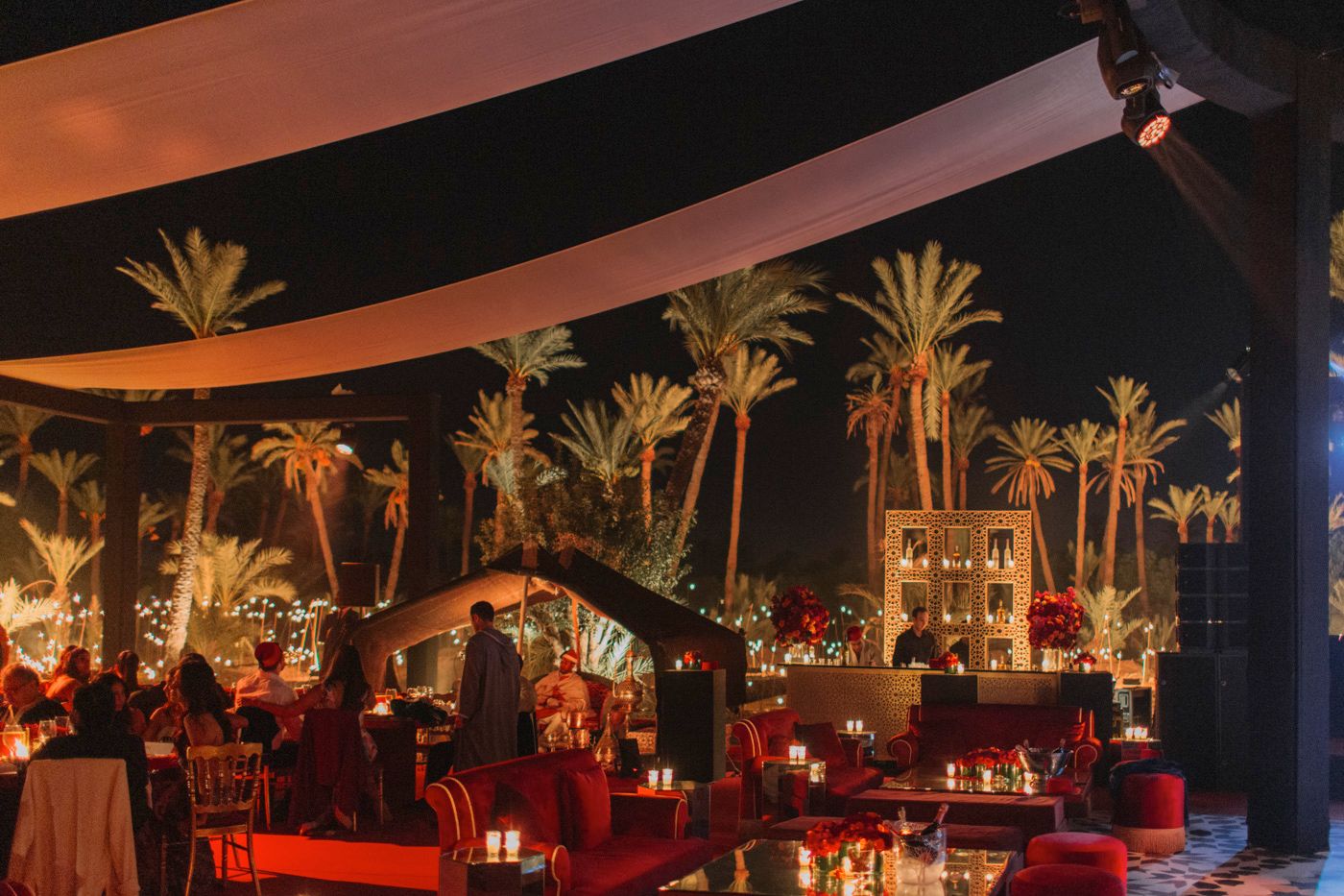 Outdoor evening event space with palm trees, string lights and red lounge seating under canopy.