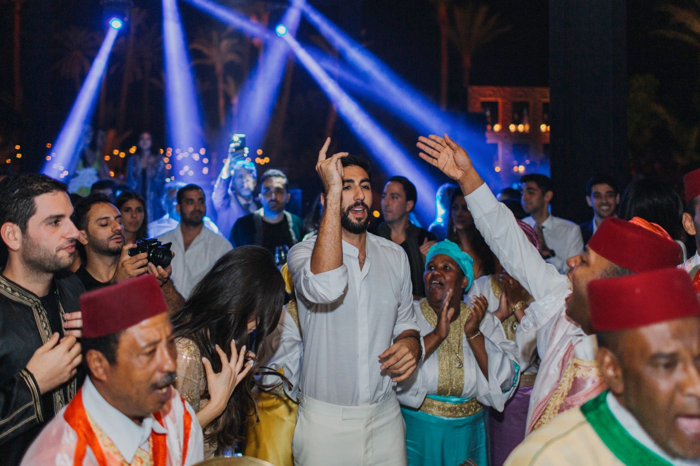 Lively party crowd wearing traditional attire dancing under blue spotlights.
