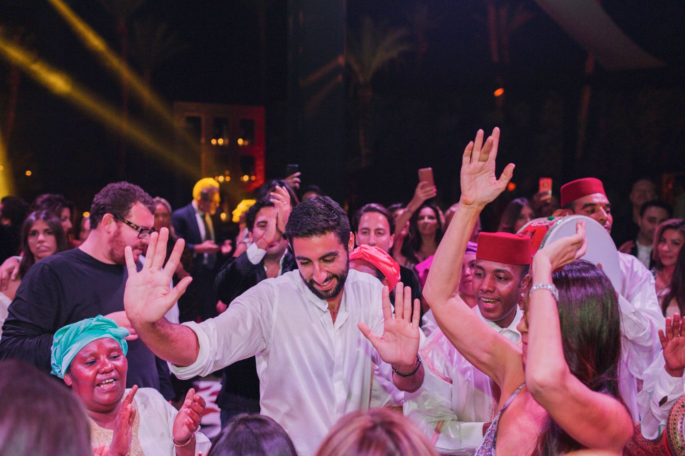 Energetic crowd dancing with raised hands under colorful party lighting.