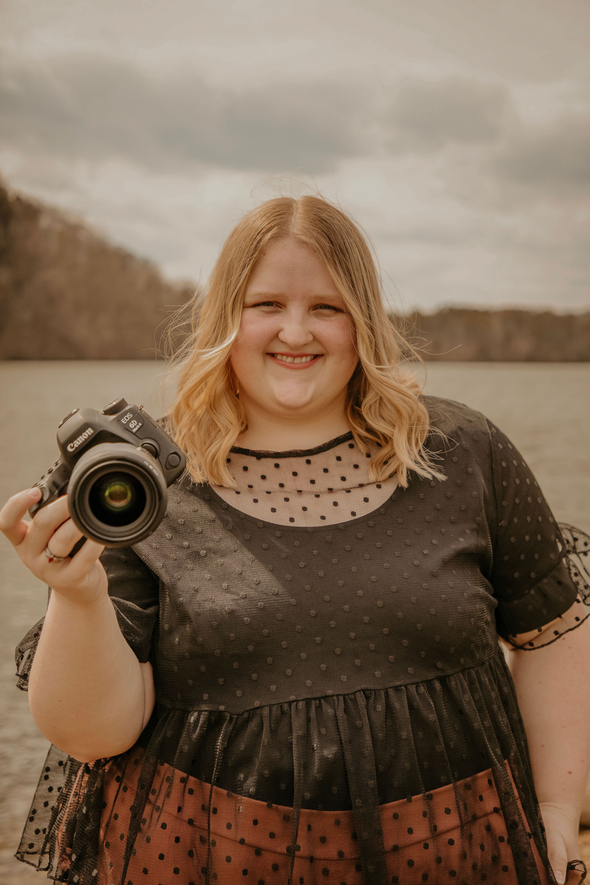 A photographer in a black polka dot dress holds a camera outdoors against an overcast lake backdrop.