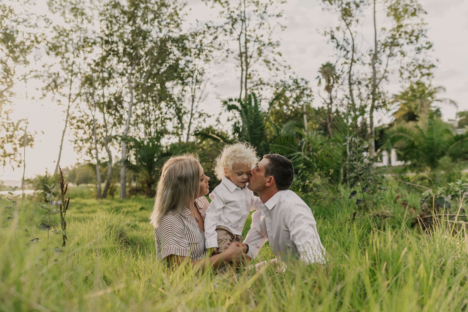 A family enjoys a picnic on a blanket while lifting their baby in a sunny park setting.