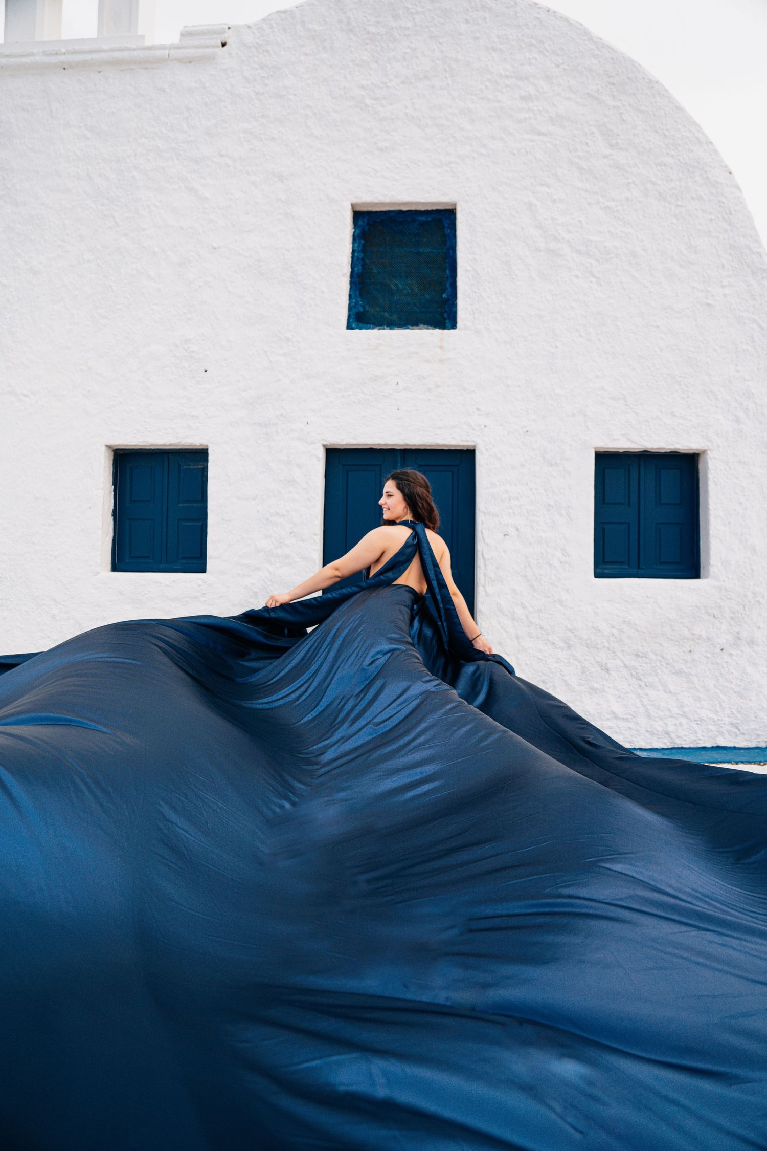 Navy blue dress billowing dramatically against white building with blue shuttered windows.