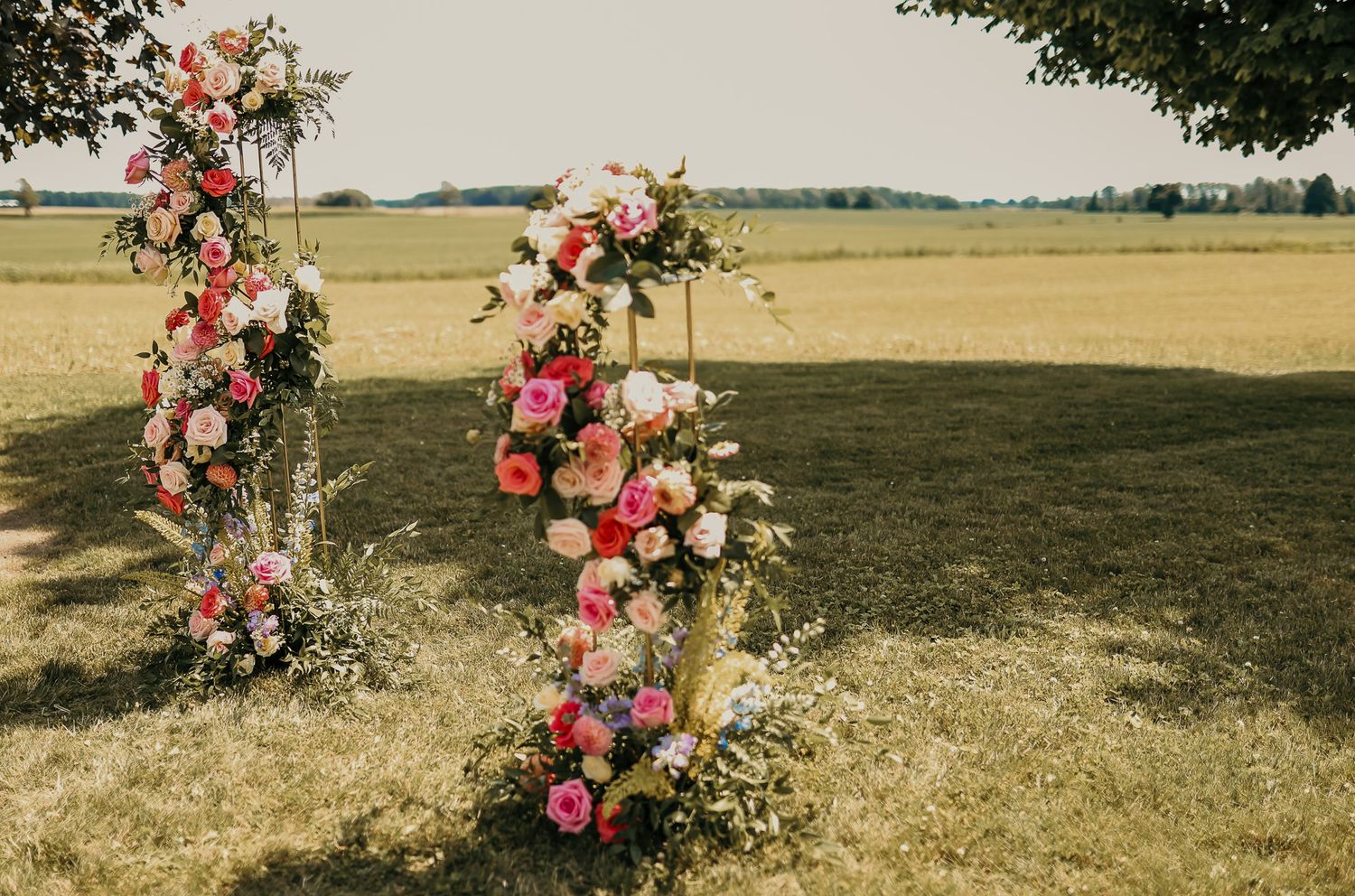 Floral wedding arch with pink and white roses stands in an open field at sunset.