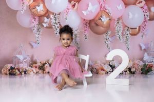 Toddler girl in pink dress sits before floral backdrop with balloons and number 2 for a second birthday photo shoot.