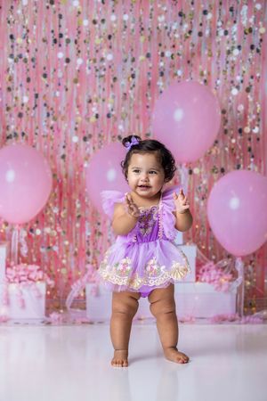 A baby girl in a purple tutu dress stands smiling at her first birthday party with pink balloons and glitter backdrop.