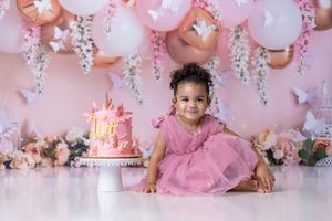 Toddler girl in pink tutu dress poses with unicorn birthday cake amid pink balloons and floral backdrop.