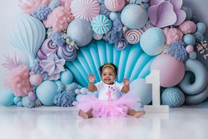Baby girl in pink tutu celebrates first birthday with colorful candy-themed backdrop featuring balloons and paper flowers.