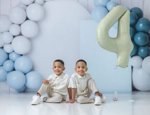 Twin boys in white outfits sit together at a 4th birthday party with blue and white balloon decorations.