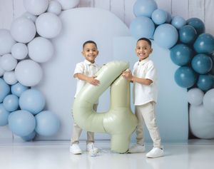 Two twin boys in white outfits hold a large number 4 balloon at a birthday party with blue and white balloon backdrop.