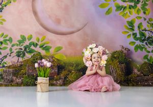 Toddler girl in pink dress and floral crown sits smiling before enchanted forest backdrop with moon and flowers.