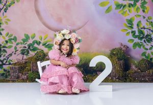 A toddler girl in a pink dress and floral crown sits beside a number 2 sign against a whimsical garden backdrop.