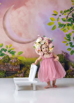 Toddler girl in pink tutu dress and floral crown smiles beside white chair against dreamy fairy tale backdrop.