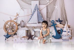 Baby boy sits among nautical decorations including anchor, ship wheel, and sailboats for a first birthday cake smash photo.