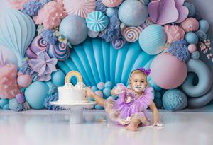 Baby girl in purple dress celebrates first birthday with white cake against colorful balloon and candy backdrop.