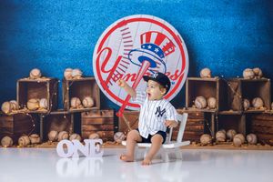 Baby in Yankees outfit sits before NY Yankees logo backdrop with baseballs, celebrating first birthday.