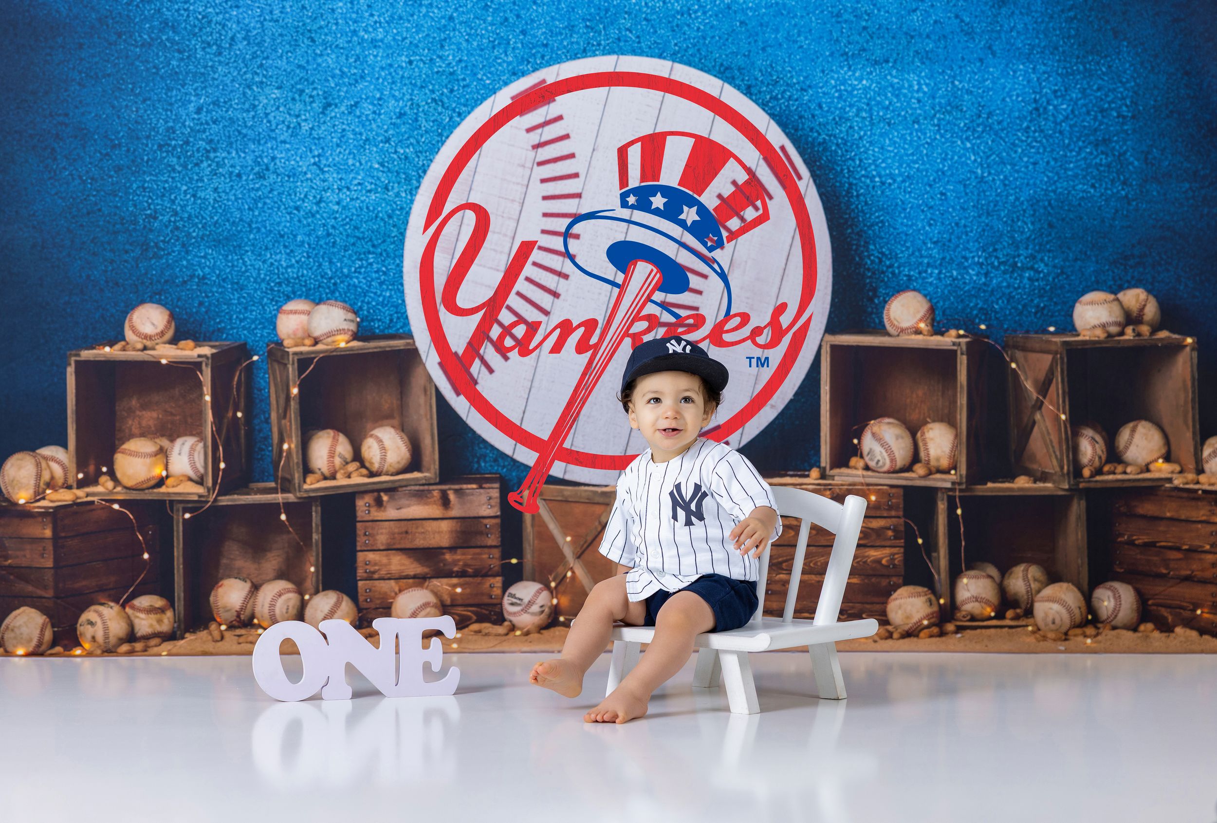 Baby in Yankees uniform sits on white chair surrounded by baseballs and crates with ONE sign for first birthday.