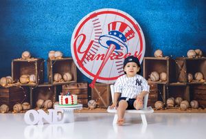 One-year-old in Yankees outfit stands by birthday cake with ONE sign and baseball crates backdrop.