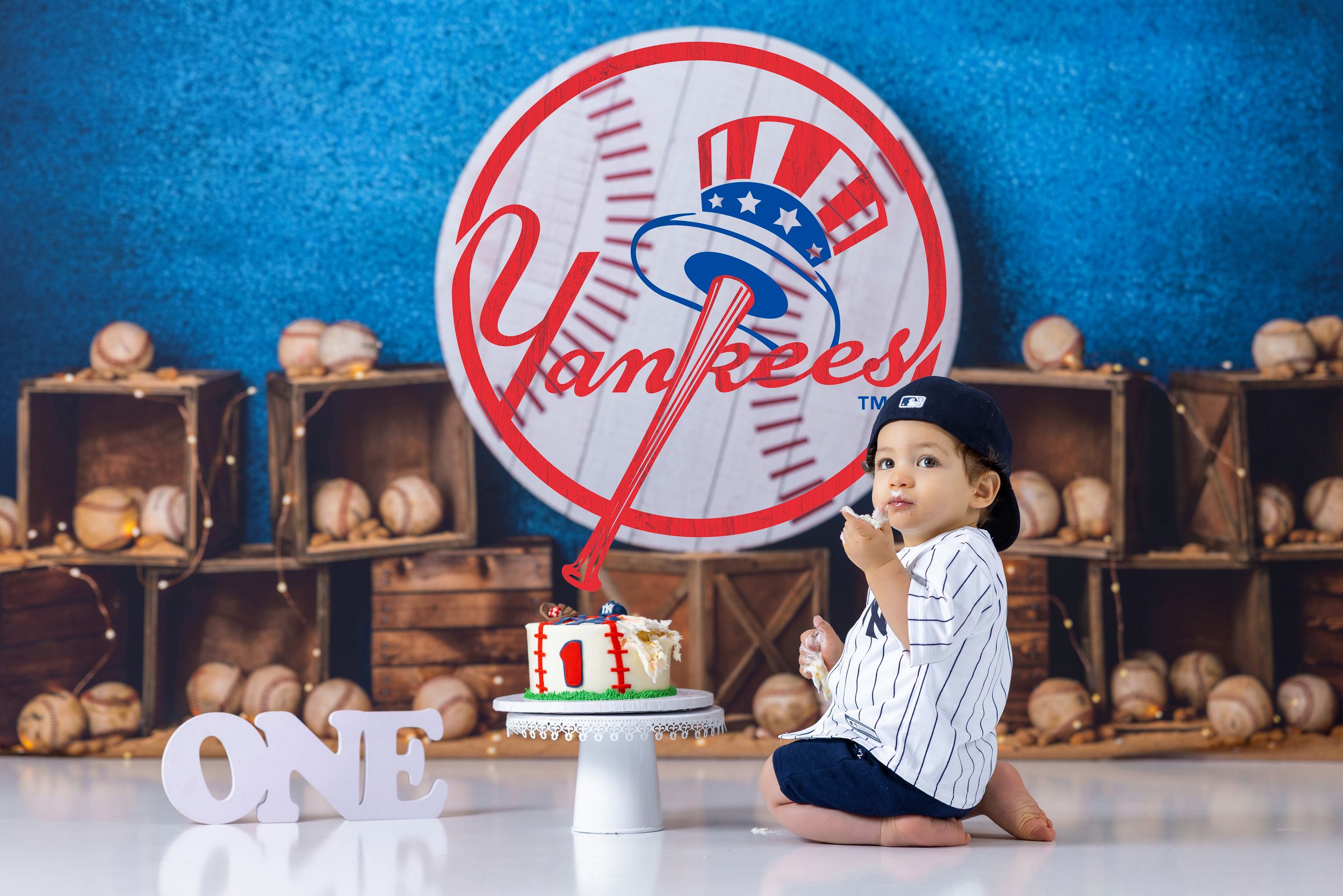 Baby boy in Yankees jersey sits beside smash cake with baseball decor and ONE sign for first birthday.