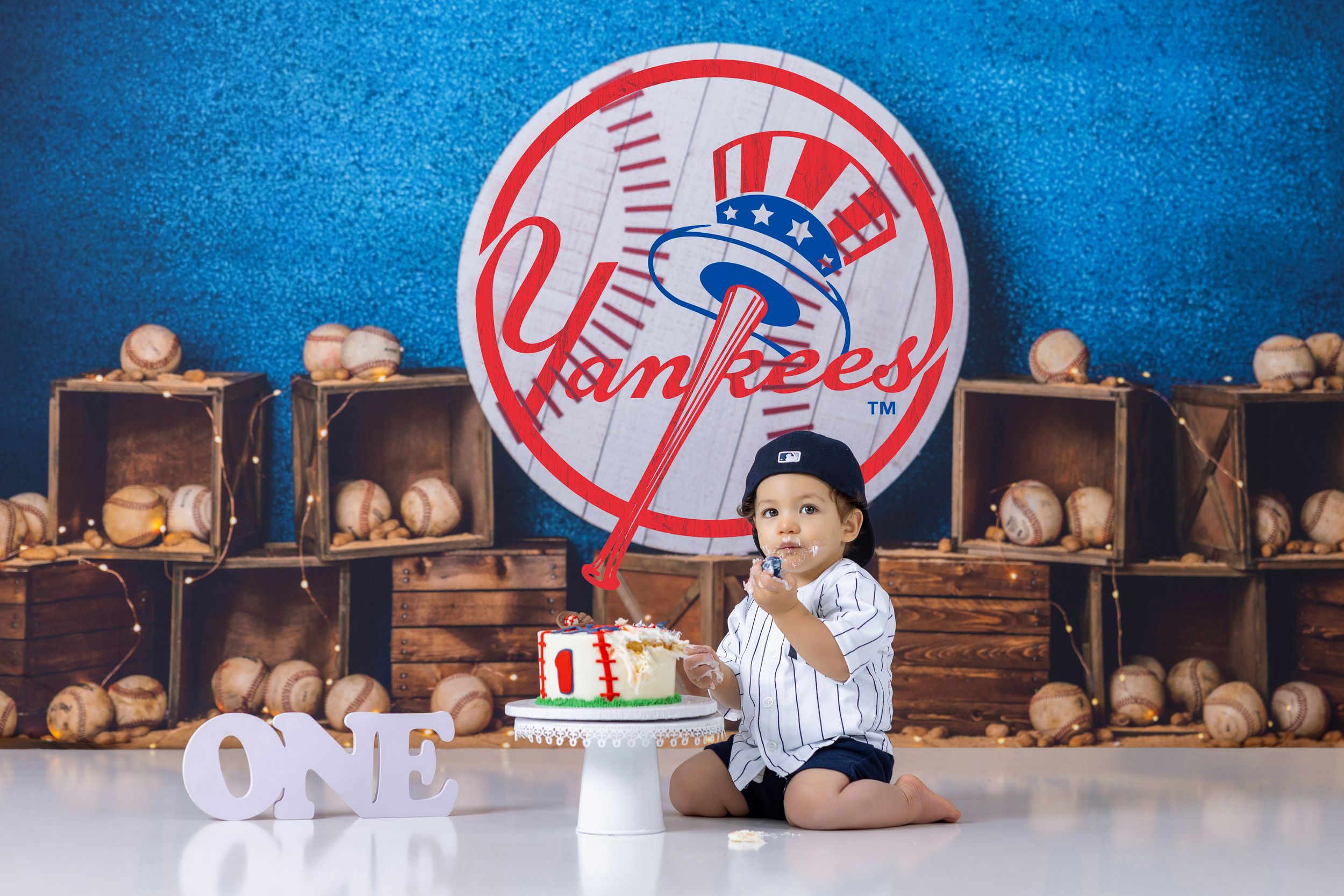 Baby boy in Yankees outfit sits beside a smash cake, surrounded by baseballs and a NY Yankees backdrop for his first birthday.