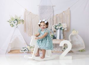 Baby girl in teal dress sits on white rocking horse with number 2, flowers, and boho teepee backdrop.