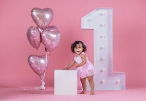 Smiling baby girl in pink tutu celebrates first birthday with heart balloons and illuminated number one on pink background.