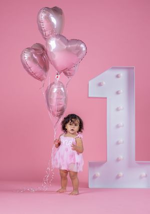 Toddler girl in pink dress holding heart balloons beside illuminated number 1 on pink background for first birthday.