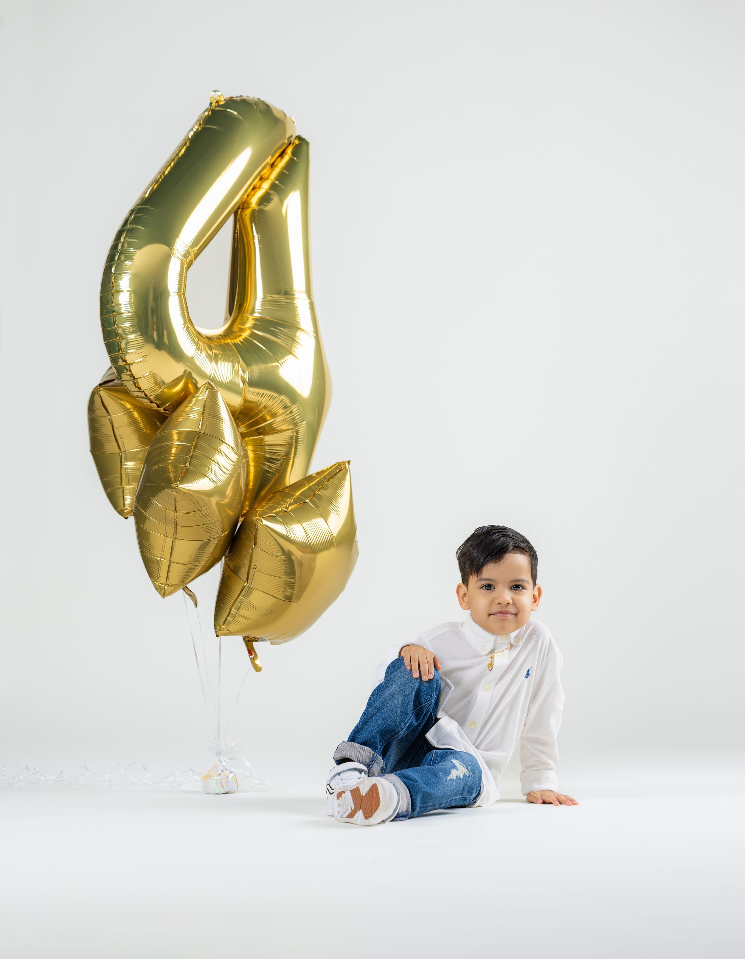 A young boy sits smiling beside gold number 4 and heart-shaped foil balloons on a white background.