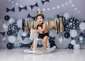 Baby in crown rides rocking horse surrounded by star balloons and bow tie decorations at a first birthday celebration.