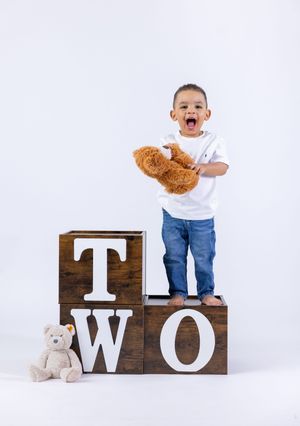 Excited toddler boy holding teddy bear standing on wooden TWO blocks for 2nd birthday photo session.