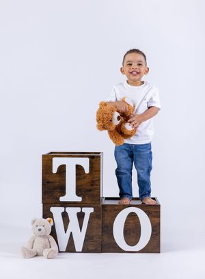 Smiling toddler boy standing on wooden TWO letter blocks holding a teddy bear, celebrating his 2nd birthday.