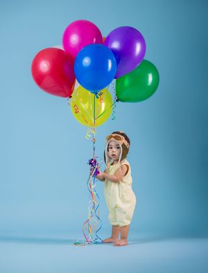 A cute toddler in a yellow romper holds colorful balloons on a blue background.