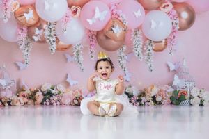 Baby girl in white tutu celebrates first birthday with pink balloons and floral backdrop in a cake smash photo session.