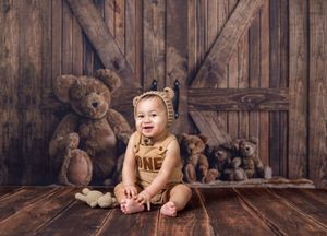 Baby in bear outfit sits smiling among teddy bears on wood floor with rustic barn door backdrop.