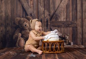 Smiling baby in bear bonnet and romper playing with basket surrounded by teddy bears on rustic wood floor.