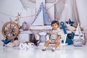 Baby boy sits among nautical decor including anchor, ship wheel, and sailboats with ONE birthday sign.