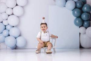 A baby boy in suspenders sits on a small white chair surrounded by blue and white balloons at his first birthday.