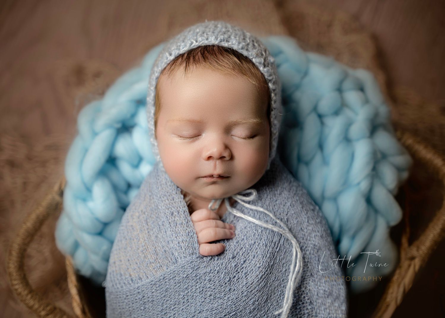 Newborn baby sleeping peacefully in a basket, wrapped in soft blue knit fabric with a matching bonnet.