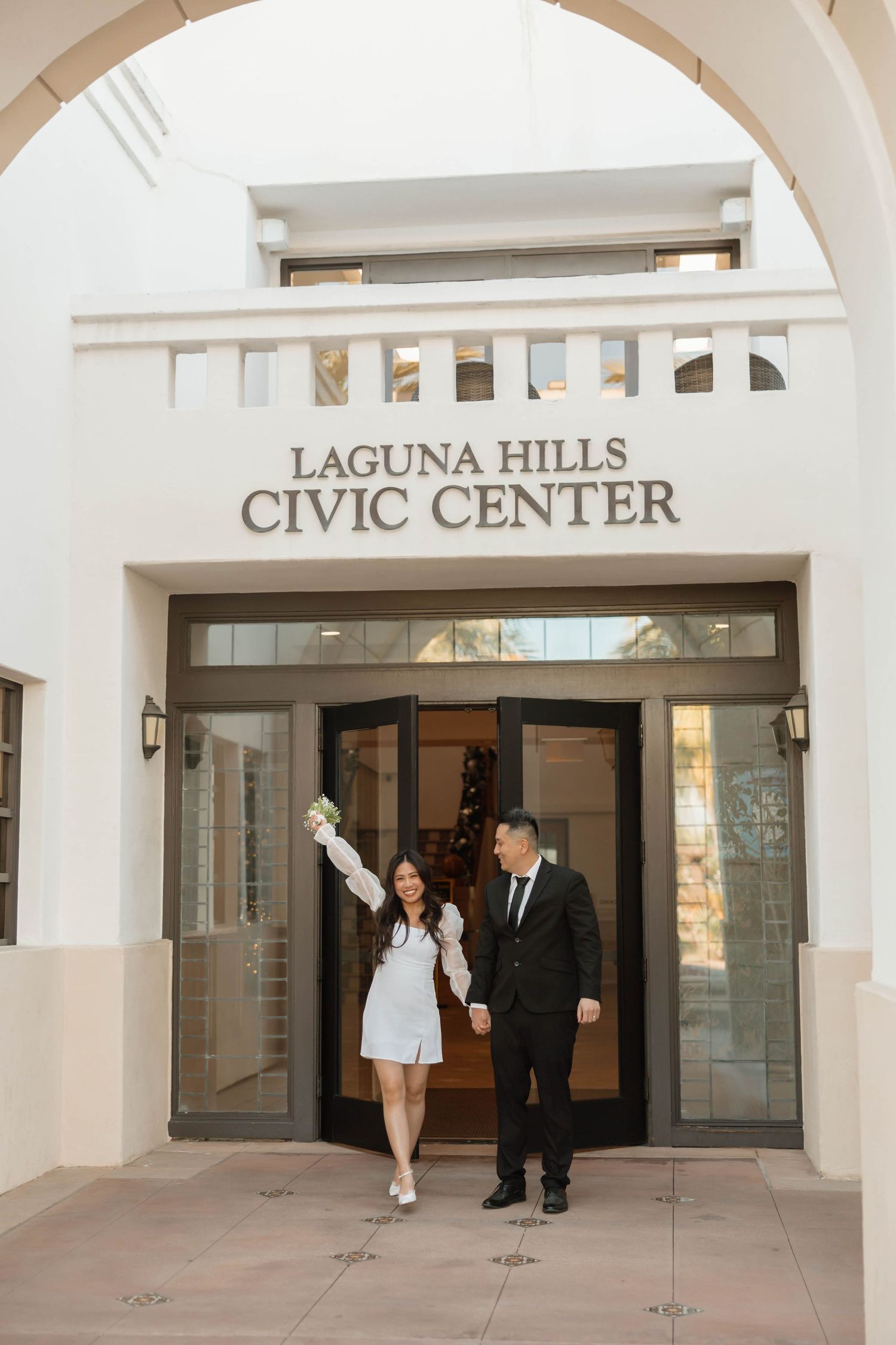 A bride and groom walk through the entrance of Laguna Hills Civic Center with white arches and columns.