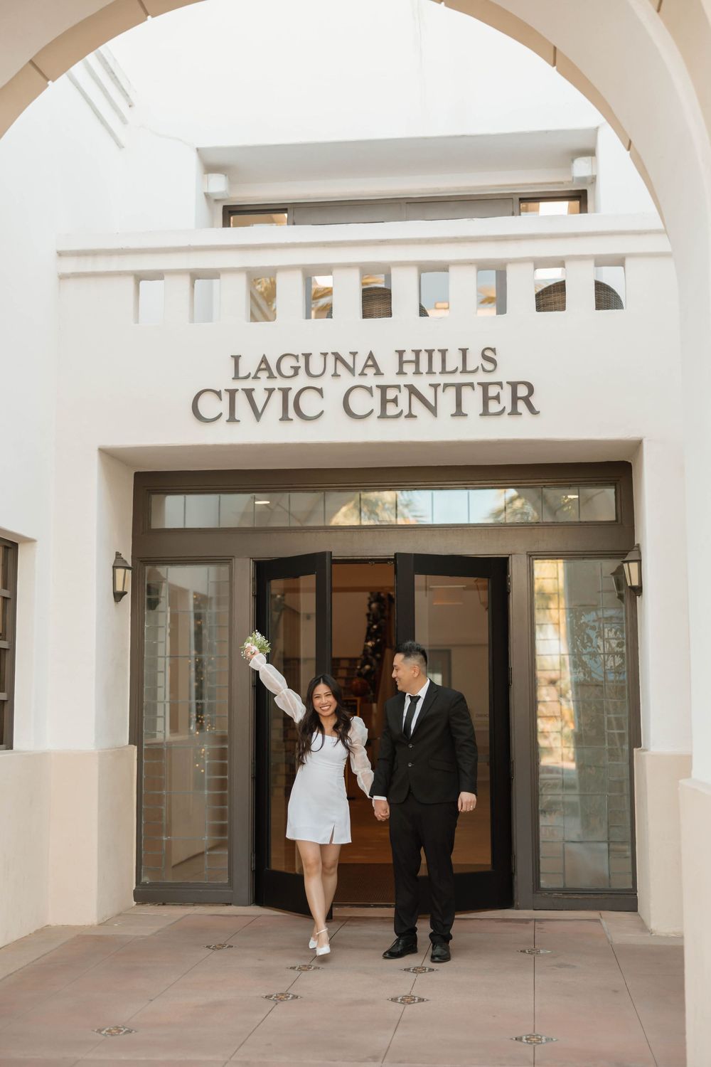A bride and groom walk through the entrance of Laguna Hills Civic Center with white arches and columns.