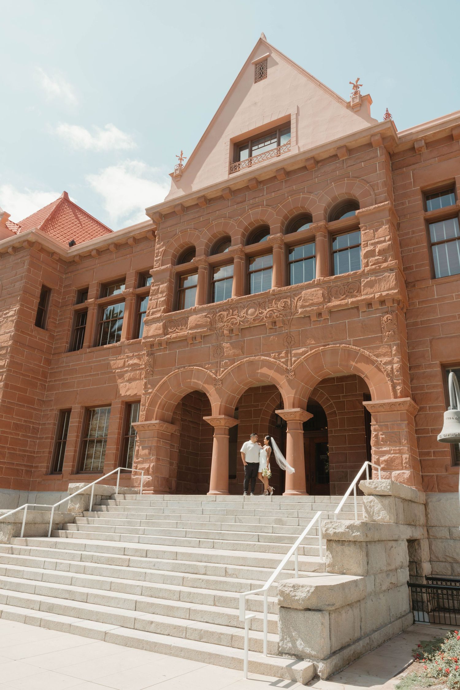 Couple walking up grand stone steps of ornate red brick building with arched windows under blue sky.