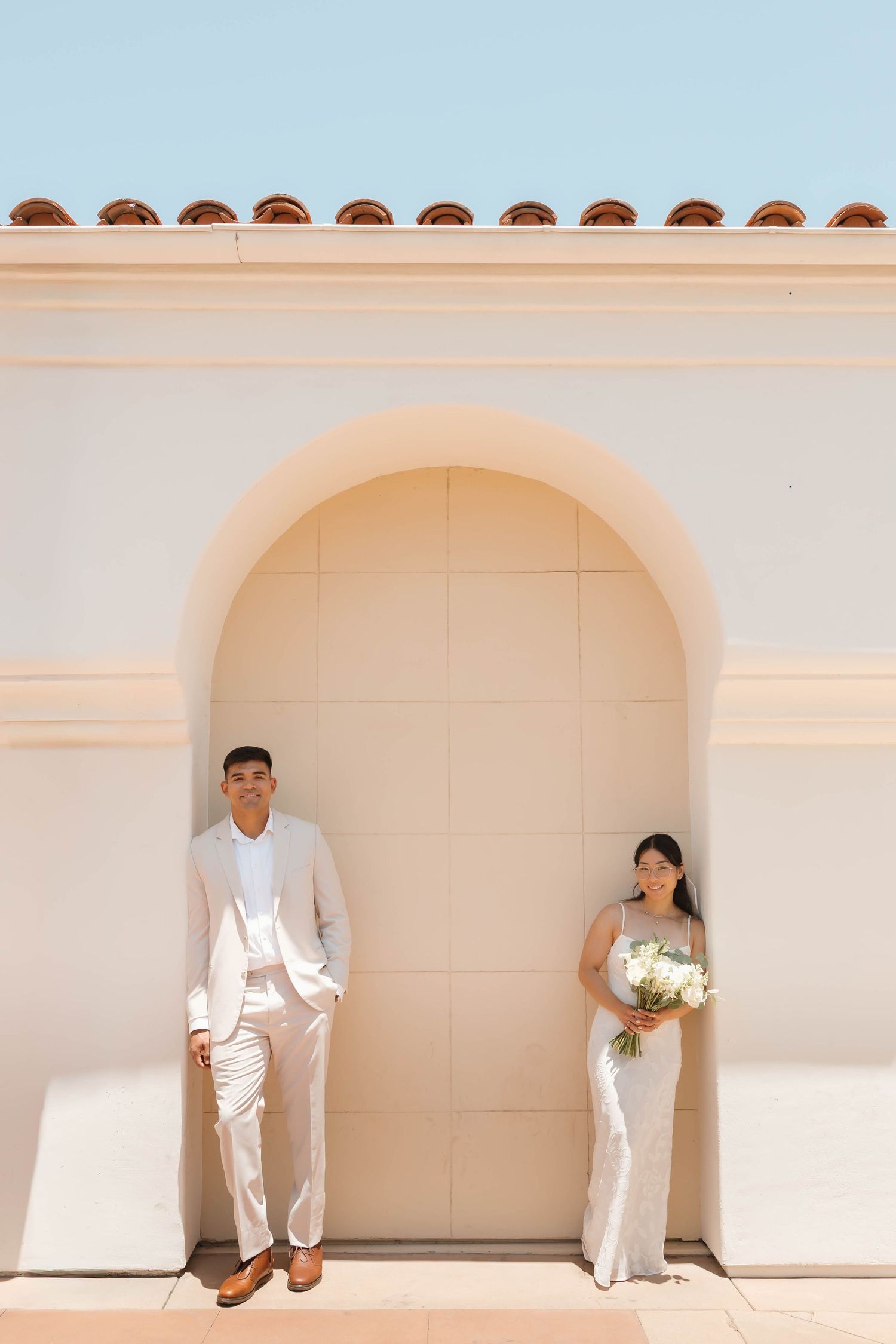 Bride and groom pose on either side of an arched doorway at an elegant cream-colored building on their wedding day.
